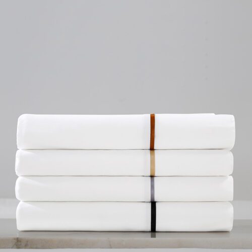 A stack of four folded luxury white hotel sheets, each featuring a different colored contrast piping: copper, gold, silver, and black.