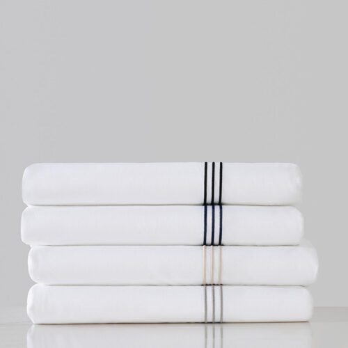 A stack of four folded luxury white hotel sheets, each featuring triple-line embroidery in black, navy, champagne, and silver.