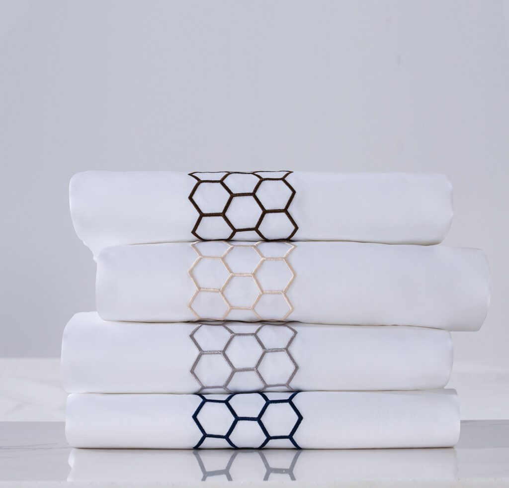 A stack of four folded luxury white hotel sheets, each featuring a different colored honeycomb hexagon embroidery: coffee, champagne, silver, and navy.
