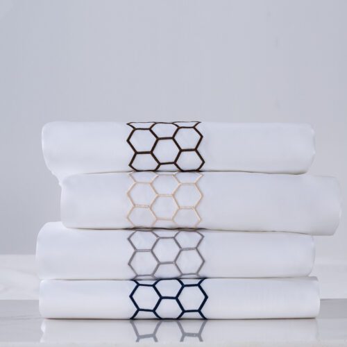 A stack of four folded luxury white hotel sheets, each featuring a different colored honeycomb hexagon embroidery: coffee, champagne, silver, and navy.