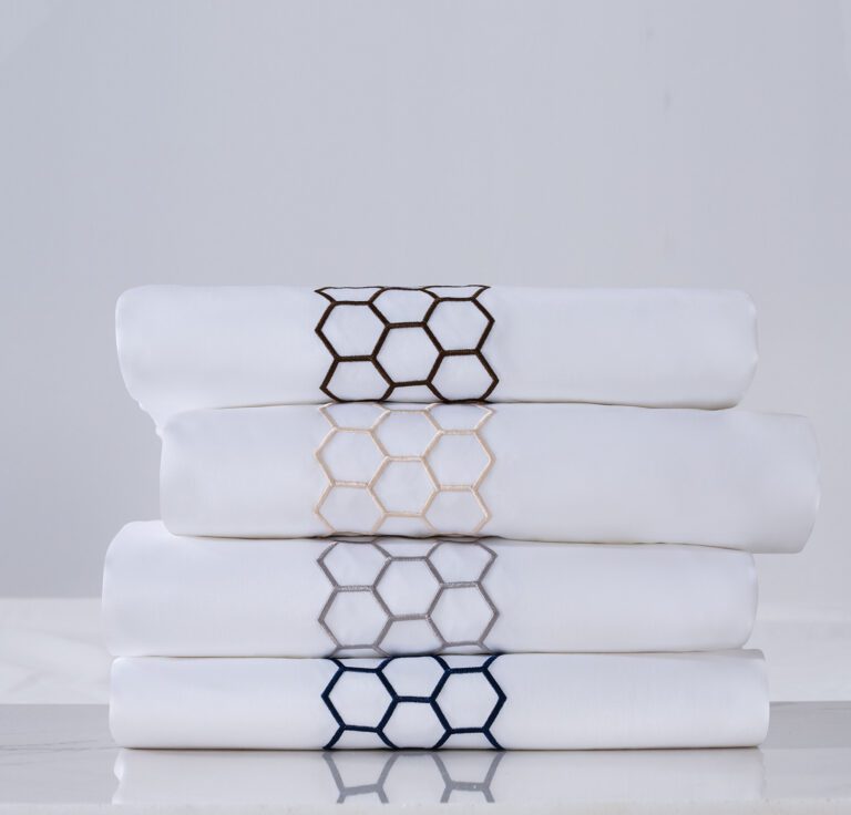 A stack of four folded luxury white hotel sheets, each featuring a different colored honeycomb hexagon embroidery: coffee, champagne, silver, and navy.
