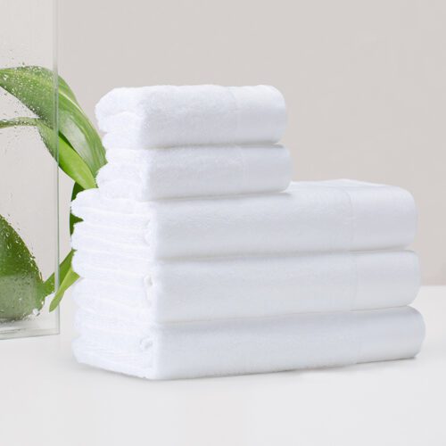 A full set of stacked white plush cotton towels in various sizes, including bath towels, hand towels, and washcloths, featuring textured dobby borders.
