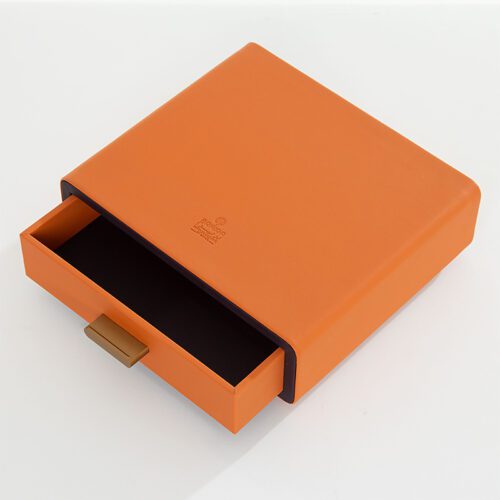 A Braira branded square vibrant orange faux leather drawer storage box with a deep purple interior and a gold-finish metal pull handle.