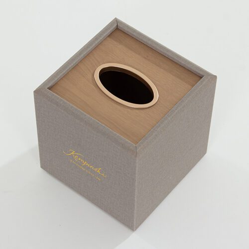 A Kempinski branded grey textured faux leather square tissue box cover with a tan wood-grain top and oval dispensing hole.