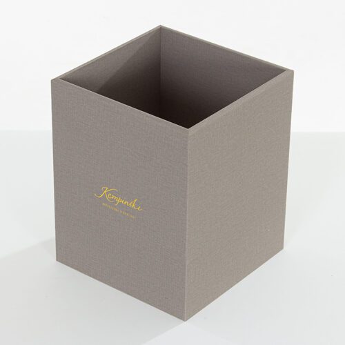 A professional shot of a Kempinski branded square grey faux leather waste basket featuring a gold foil script logo.