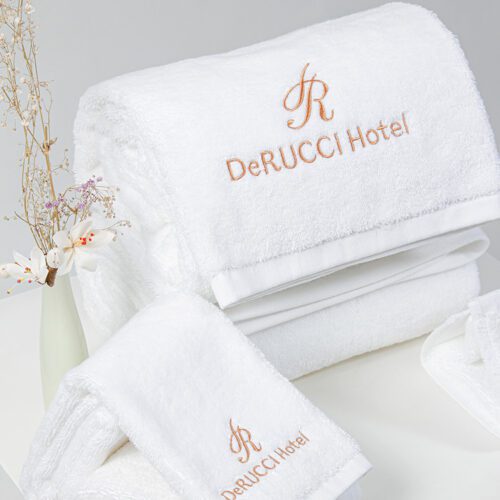 A set of folded white plush cotton towels featuring the DeRUCCI Hotel logo and "R" crest embroidered in rose gold.