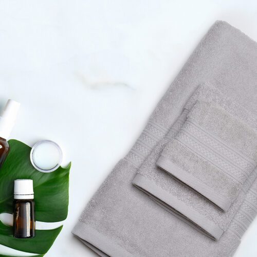 A set of folded light grey plush cotton towels with decorative dobby borders, styled next to a green leaf and aromatherapy oil bottle.