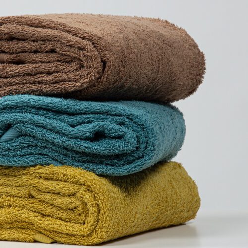 A stack of three thick, plush cotton towels in coffee brown, deep teal, and mustard yellow showing their soft texture.