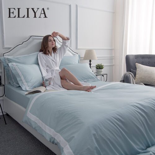 A modern luxury bed featuring a woman in a robe relaxing on a light teal duvet cover with a crisp white frame border and matching Oxford pillowcases.