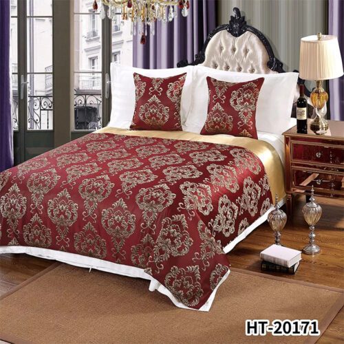 A luxury hotel bed with white linens featuring a deep red bedspread with golden Damascus floral patterns and a gold satin border.