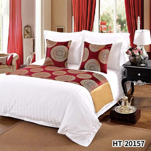A luxury hotel bed with white striped linens featuring a red bed runner with golden mandala circles and a gold satin accent.