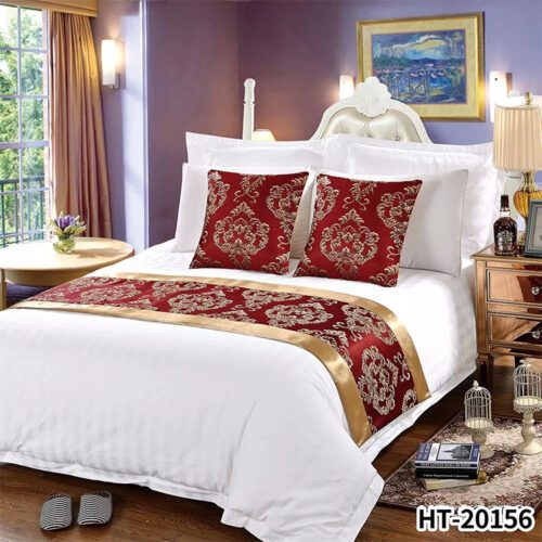A luxury hotel bed with white striped linens featuring a wine red damask bed runner and two matching pillows with gold satin borders.