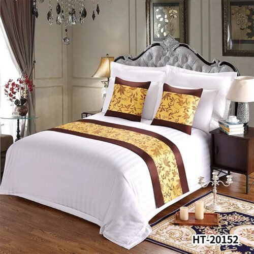 A classic hotel bed with white striped linens featuring a golden-yellow leaf patterned runner and pillows with brown satin borders.