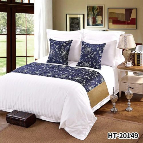 A modern hotel bed with white striped linens featuring a navy blue runner with silver firework patterns and a gold accent panel.