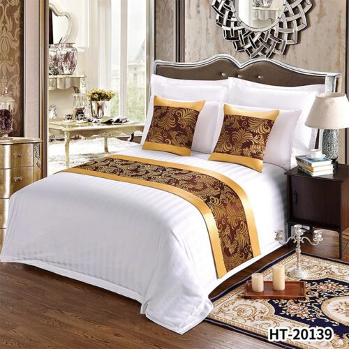 A luxury hotel bed with white striped linens featuring a golden bronze damask bed runner and two matching pillows with gold satin borders.
