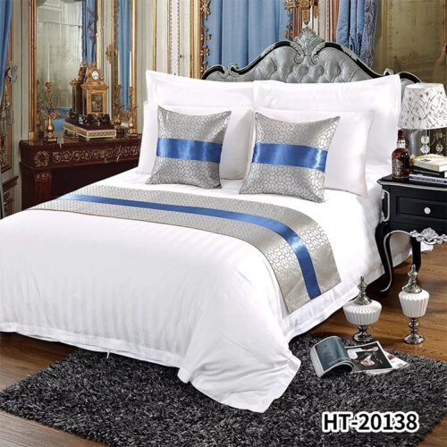 A luxury hotel bed with white striped linens featuring a silver geometric jacquard runner and matching pillows with a blue satin stripe.