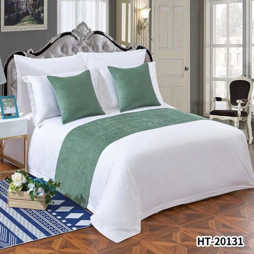 A minimalist hotel bed with white linens featuring a sage green velvet runner and two matching square pillows.
