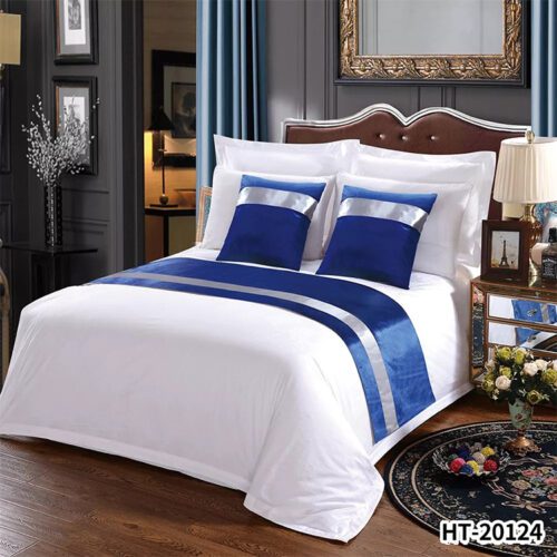 A luxury hotel bed with white linens featuring a royal blue velvet runner and matching pillows with silver satin stripes.