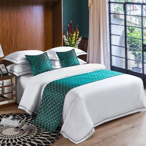 A luxury hotel bed with white linens featuring an emerald green bed scarf with peacock feather patterns and matching pillows.