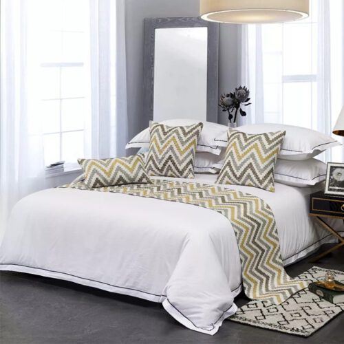 A professional hotel bed with white linens featuring an earth tone chevron bed scarf and matching pillows in a bright room.