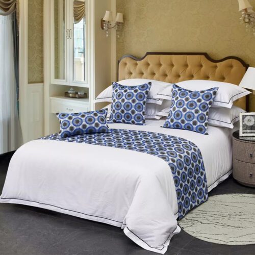 A professional hotel bed with white linens featuring a silver grey bed scarf with a black Greek key border and matching pillows.