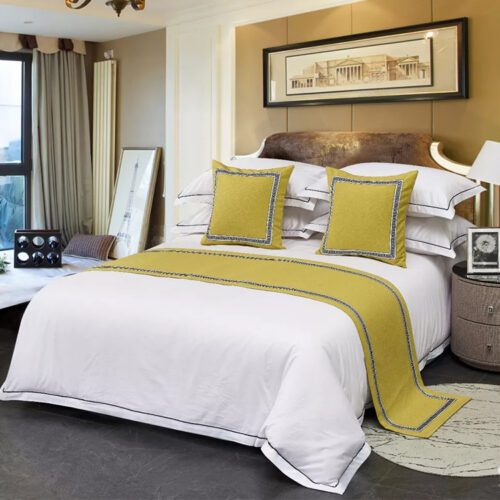A professional hotel bed with white linens featuring a ginger yellow bed scarf with a black Greek key border and matching pillows.