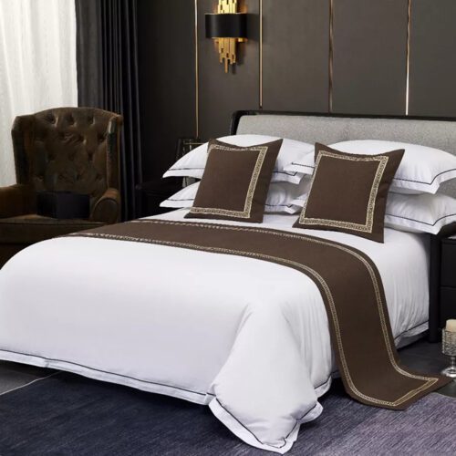 A professional hotel bed with white linens featuring a deep coffee brown bed scarf with a gold Greek key border and matching pillows.