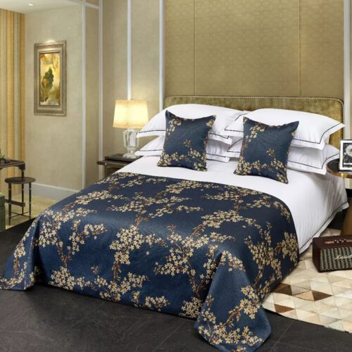 A luxury hotel bed with white linens featuring a navy blue bedspread with golden plum blossom patterns and matching pillows.