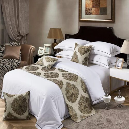 A traditional luxury hotel bed with white linens featuring a champagne damask floral bed scarf and matching pillows.