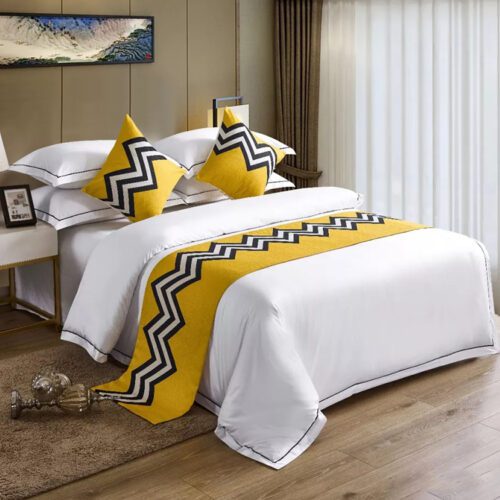 A modern hotel bed with white linens featuring a bright yellow bedspread with black and white zigzag patterns and matching pillows.