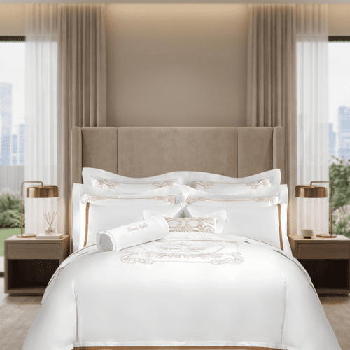 A luxury hotel bed featuring white linens with intricate rose gold embroidery on the duvet and pillows in a modern suite.