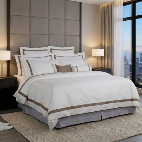 A fully made luxury hotel bed featuring white linens with a double-line brown embroidered border in a modern executive suite.