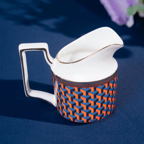 A luxury porcelain milk pitcher featuring a vibrant orange and blue 3D geometric pattern and a gold-rimmed spout and handle.