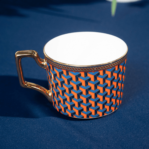 A luxury porcelain teacup featuring a vibrant orange and blue 3D geometric pattern and an ornate gold handle.