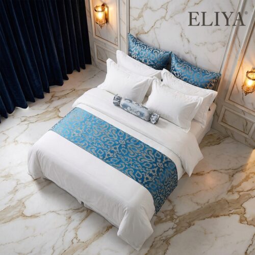 An overhead view of a luxury white hotel bed layered with a blue and gold damask jacquard bed runner and matching decorative pillows.