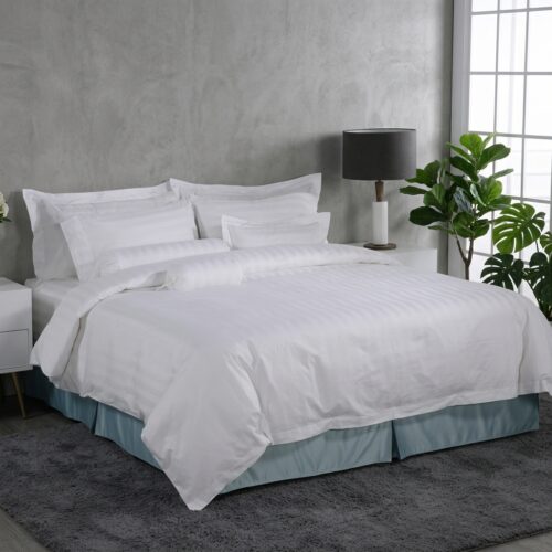 A luxury hotel bed featuring white satin-striped linens, matching bolster pillows, and a pleated coastal blue bed skirt against a grey wall.