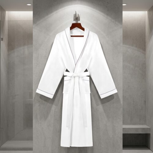 A luxury white hotel bathrobe with grey piping trim hanging on a wooden hanger against a modern grey concrete wall.