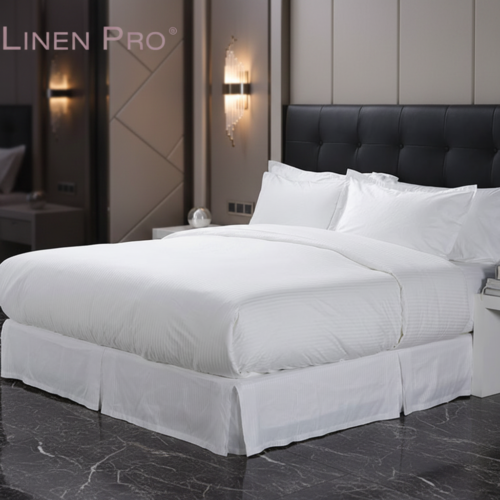 A luxury white hotel bed featuring satin stripe linens and a tailored white pleated bed skirt against a black tufted headboard.