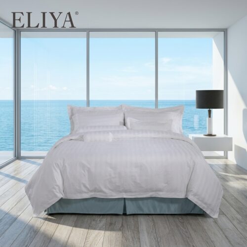 A luxury hotel bed with white satin-striped linens, a matching bolster pillow, and a coastal blue bed skirt overlooking the ocean.