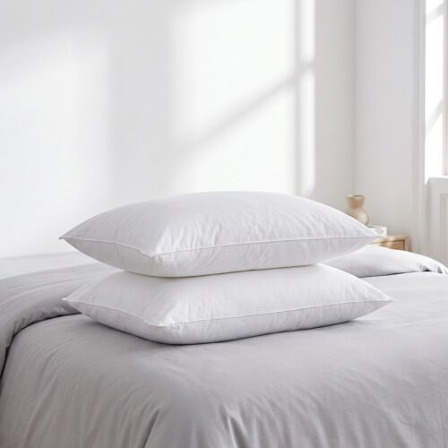 A neat stack of two white down-alternative hotel pillows on a bed with light grey linens.