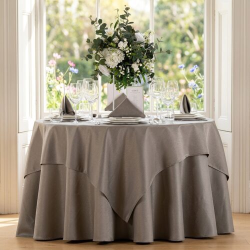 A round banquet table set for a wedding with champagne-colored linens, a layered square overlay, and a large floral centerpiece.