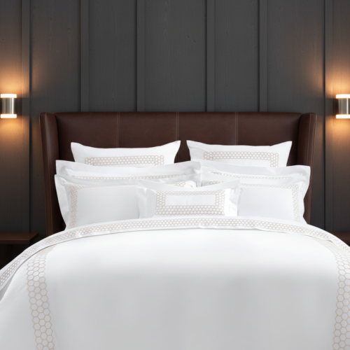 A luxury hotel bed with white linens featuring a geometric honeycombed embroidery in champagne gold against a brown leather headboard.