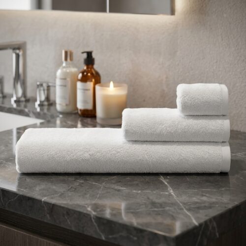A neatly folded stack of luxury white cotton hotel towels arranged on a grey marble vanity countertop next to a lit scented candle.