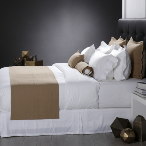 A luxury white hotel bed layered with a solid camel brown bed runner and matching brown accent pillows against a dark headboard.