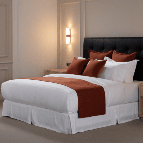 A luxury hotel bed with white linens, a white pleated bed skirt, and sienna-colored decorative pillows and bed runner.