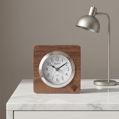 A square walnut wooden alarm clock with the ELIYA brand name on a white marble nightstand next to a brushed chrome desk lamp.