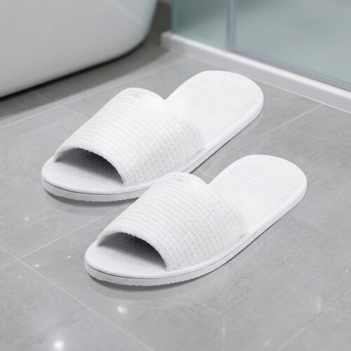 A pair of premium white open-toe waffle hotel slippers resting on a grey tiled bathroom floor next to a glass shower door.