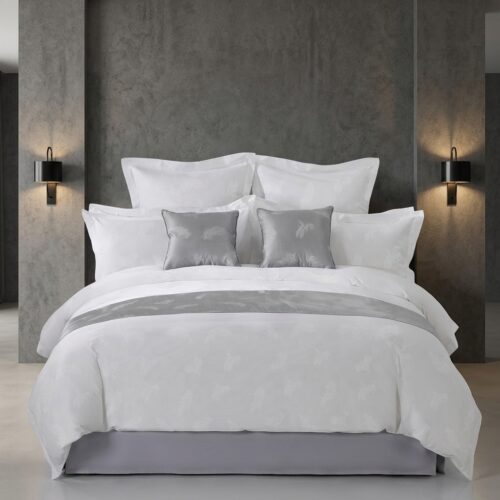 A luxury hotel bed featuring white leaf jacquard linens, a silver bed runner, and matching silver decorative pillows against a concrete-textured wall.