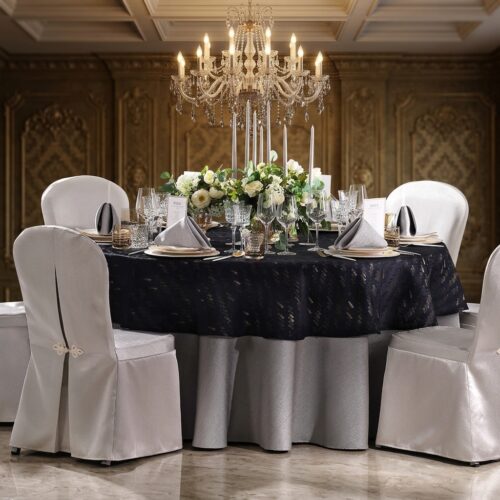 Luxury black jacquard round tablecloth with gold floral pattern styled with white chair covers, crystal candelabra, and white floral centerpiece in an elegant ballroom
