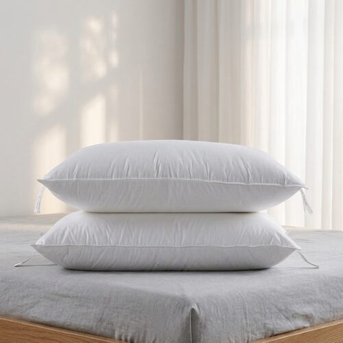 A stack of two plush white hotel pillows with double-stitched piping and corner tags resting on a grey linen bed.